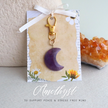 Amethyst moon-shaped bagcharm on a decorative card with a white ribbon, set against a neutral background.