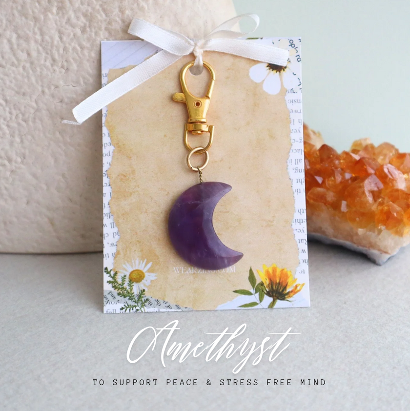 Amethyst moon-shaped bagcharm on a decorative card with a white ribbon, set against a neutral background.